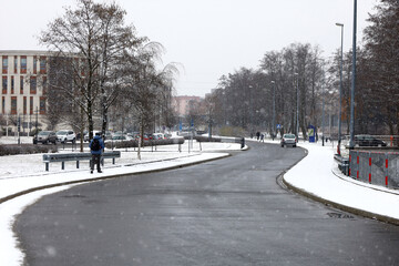 Fototapeta na wymiar Empty road in winter on background, beginning of snowfall, snowflakes defocused and blurred in foreground. Pedestrian on sidewalk, cars in the distance, not recognizable.