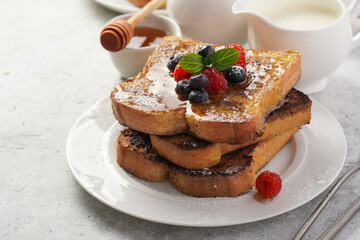 Several pieces of french toast - white wheat bread soaked in egg, milk and sugar, fried on a pan - stacked on white plate with fresh raspberries, blueberries, honey, mint on light grey background