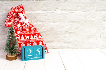 Fototapeta na wymiar Red gift bag, decorative Christmas tree and blue wooden perpetual calendar of cubes on white brick wall background. Top view, flat lay with copy space, banner, header, New Year background