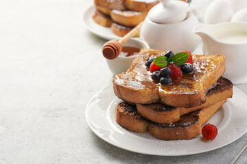 Several pieces of french toast - white wheat bread soaked in egg, milk and sugar, fried on a pan - stacked on white plate with fresh raspberries, blueberries, honey, mint on light grey background