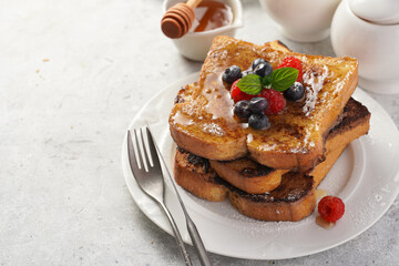 Several pieces of french toast - white wheat bread soaked in egg, milk and sugar, fried on a pan - stacked on white plate with fresh raspberries, blueberries, honey, mint on light grey background