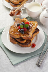 Several pieces of french toast - white wheat bread soaked in egg, milk and sugar, fried on a pan - stacked on white plate with fresh raspberries, blueberries, honey, mint on light grey background