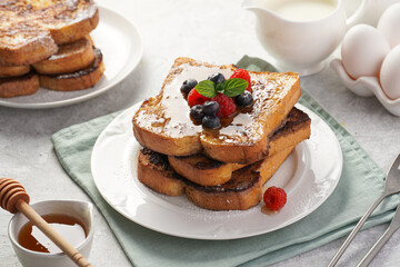 Several pieces of french toast - white wheat bread soaked in egg, milk and sugar, fried on a pan - stacked on white plate with fresh raspberries, blueberries, honey, mint on light grey background