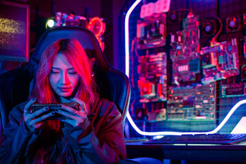 Gamer playing online game application on mobile phone wear gaming headphones, Asian woman live stream she play video game via smartphone at home neon lights living room