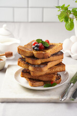 Several pieces of french toast - white wheat bread soaked in egg, milk and sugar, fried on a pan - stacked on white plate with fresh raspberries, blueberries, honey, mint on light grey background