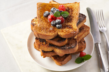 Several pieces of french toast - white wheat bread soaked in egg, milk and sugar, fried on a pan - stacked on white plate with fresh raspberries, blueberries, honey, mint on light grey background
