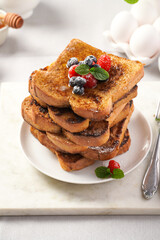 Several pieces of french toast - white wheat bread soaked in egg, milk and sugar, fried on a pan - stacked on white plate with fresh raspberries, blueberries, honey, mint on light grey background