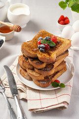 Several pieces of french toast - white wheat bread soaked in egg, milk and sugar, fried on a pan - stacked on white plate with fresh raspberries, blueberries, honey, mint on light grey background