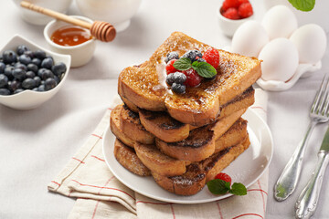 Several pieces of french toast - white wheat bread soaked in egg, milk and sugar, fried on a pan - stacked on white plate with fresh raspberries, blueberries, honey, mint on light grey background