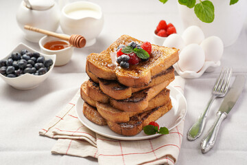 Several pieces of french toast - white wheat bread soaked in egg, milk and sugar, fried on a pan - stacked on white plate with fresh raspberries, blueberries, honey, mint on light grey background