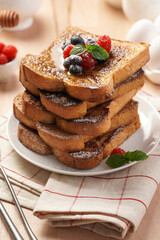 Several pieces of french toast - white wheat bread soaked in egg, milk and sugar, fried on a pan - stacked on white plate with fresh raspberries, blueberries, honey, mint on wooden background