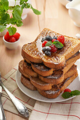 Several pieces of french toast - white wheat bread soaked in egg, milk and sugar, fried on a pan - stacked on white plate with fresh raspberries, blueberries, honey, mint on wooden background