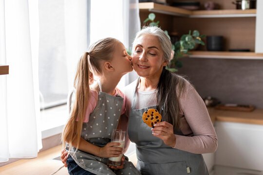 Smiling caucasian little granddaughter kisses elderly grandmother on cheek with cookies, enjoy baking