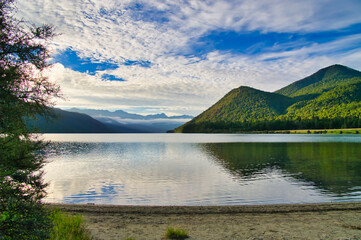 Lake Rotoroa, Nelson Lakes National Park, New Zealand, on an early summer morning, with the low sun illuminating the green mountains and mist hanging in the valley
