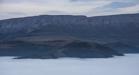 Mountain evening landscape with low clouds in the valley and a steep cliff in the background, panorama of the evening mountains