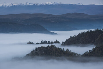 Low clouds in the mountains filled the entire valley and rocky peaks and hills with trees stick out above the clouds, mountains with snow in the background, fog between the mountains on an autumn