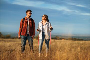 A couple is chatting and hiking in big golden yellow meadow on a sunny autumn day. They wear sunglasses to protect them from sun. The girl is holding trekking poles to help her while hiking.