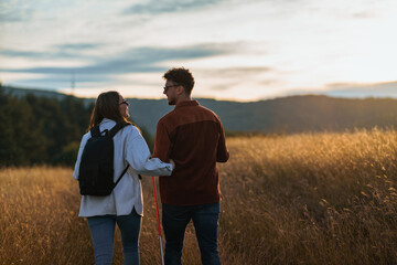 A girl is holding a boy's arm during their hike through a big golden yellow meadow on a sunset, talking to each other and wearing sunglasses.