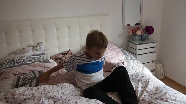A 10-year-old Caucasian boy does somersaults on a family bed. The brother jumps, somersaults and rolls over in the air. Somersault jump. Slow motion. Flying pillow. Morning in the children's bedroom