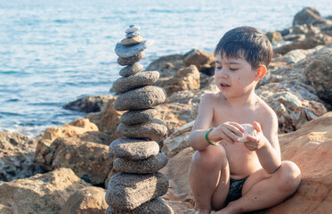 kid boy sitting on big rock seashore coast line playing with stones building pyramid.pebbles one over another zen wellbeing yoga relax meditation spa concept.water waves hit beach summer vacation