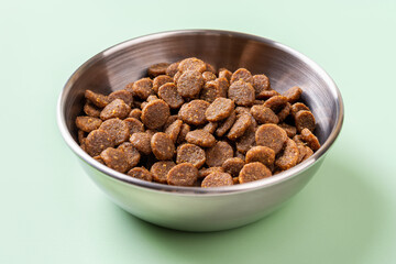 Dry pet food in a metal bowl over green background. Gluten free dry protein kibbles for cats in a stainless steel feeding bowl closeup. Complete dry food for domestic animals concept.