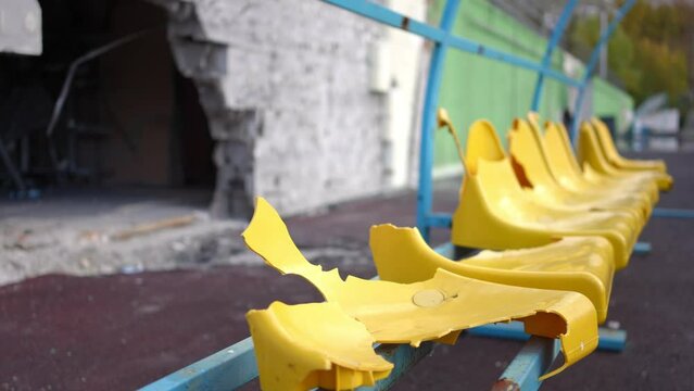 Side view row of yellow damaged spectator seats outdoors at background of wall with bomb hole. Live camera moves left and right outdoors at destroyed children's stadium in Kiev suburb Ukraine
