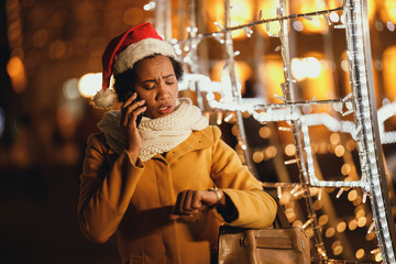 Black Woman Looking At Her Watch And Using Smartphone At Christmas Time In The City