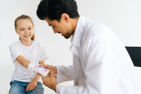 Friendly pediatrician male in uniform examining smiling little girl patient with broken hand wrapped in plaster bandage at checkup meeting, on white isolated background in professional studio.