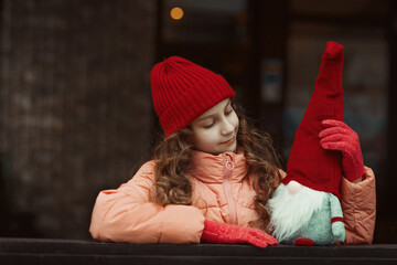 Pretty dreamy young girl teenager in red hat and pink coat looking at traditional christmas gnome toy in big red cap with long beard, festive outdoor seasonal new year portrait, dark brown background