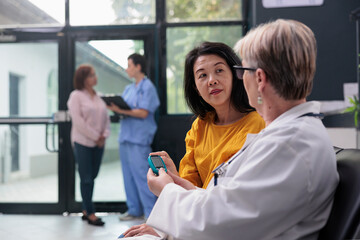 Obraz premium Senior medic consulting asian patient with glucometer instrument, measuring insulin and glucose level from blood sample. Doctor and woman doing diabetic control at checkup visit appointment.