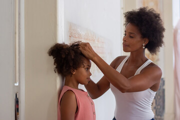 African American mom measuring daughters height. Girl in pink clothes standing near wall, young...