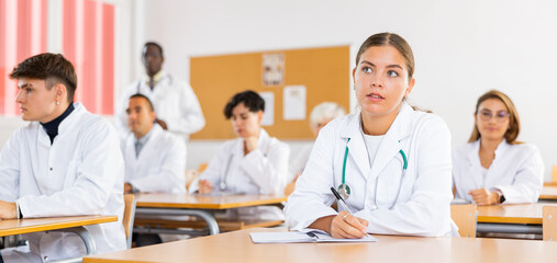 Girl medical student sitting at desk in classroom during lecture in medical college with group of students.