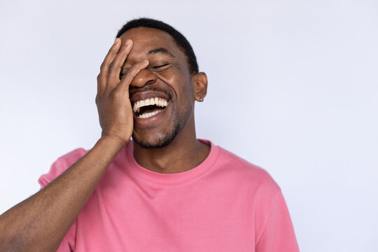 Portrait of happy African American man laughing hard. Amused young male model with short dark hair in pink T-shirt with closed eyes smiling, covering face with hand. Humor, happiness concept