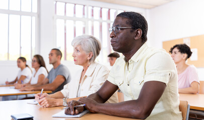 Adult african-american man sitting at desk in classroom during foreign language course, listening to lecture carefully.