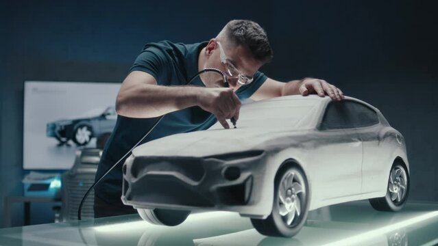 Senior car designer and developer works on a prototype car sculpture made of clay. Doing the final touch ups using the ponited end of the milling machine. A high tech innovative office.