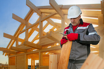 Builder worker. Man with ruler measures board. Worker builds house out of wood. Guy in form of builder. Builder man near frame of wooden house. Concept of checking building materials for quality