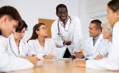 Fototapeta premium Group of medical workers in white coats sitting at same desk during professional training course, listening to experienced african american lecturer..