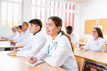 Interested young female student in white coat listening to lecture and taking notes in classroom...