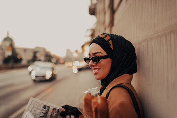 A modern Muslim woman with a hijab walks around the city while carrying a newspaper and bread in her hand