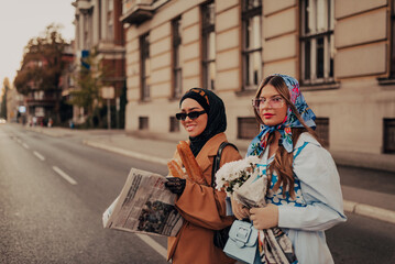 A modern European woman and a Muslim woman with a hijab walking the streets of the city dressed in clothes from the 19th century while carrying newspapers, flowers and bread in their hands.