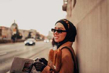 A modern Muslim woman with a hijab walks around the city while carrying a newspaper and bread in...