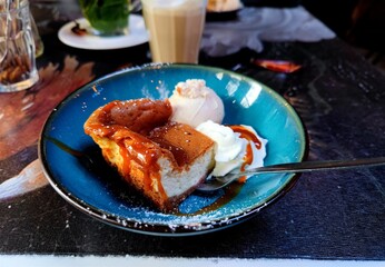 Delicious cheesecake with caramel syrup, a dollop of whipped cream and hazelnut walnut ice cream in a blue bowl on a restaurant table. There are no persons or trademarks in the shot.