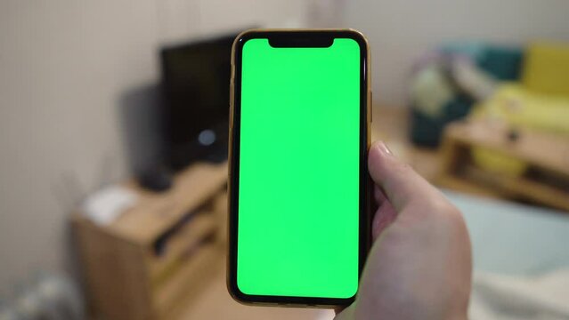 Use green screen for copy space closeup. Chroma key mock-up on smartphone in hand.  young man holds mobile phone iPhone and swipes photos or pictures left indoors of cozy home