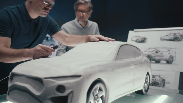 Senior automotive design engineers work on the prototype model of the car looking at the pencil sketches of the design. Using a rotary tool for final touch ups in the rake sculpture placed on a table.