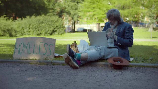 Homeless man sitting on sidewalk using laptop. Realtime