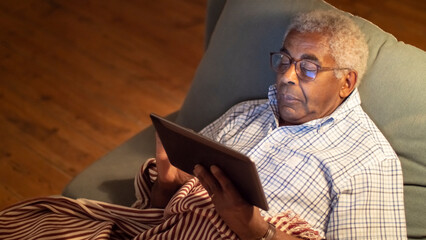 Portrait of African American man holding tablet. Elderly man lying on sofa relaxed watching news, having rest in evening. Having rest with smart devices, wi-fi communication concept