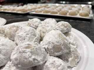 A white plate of Snowball Cookies sprinkled with powdered sugar. Baking sheet with more cookies out of focus in the background