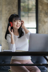 
Beautiful and cheerful Asian woman wearing headphones in video conferences and online classes on the Internet and enjoying listening to music happily on her work laptop during work breaks.