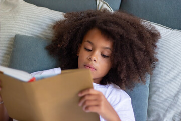 Focused African American girl reading in bed. Child with curly hair lying in bed, looking at book...