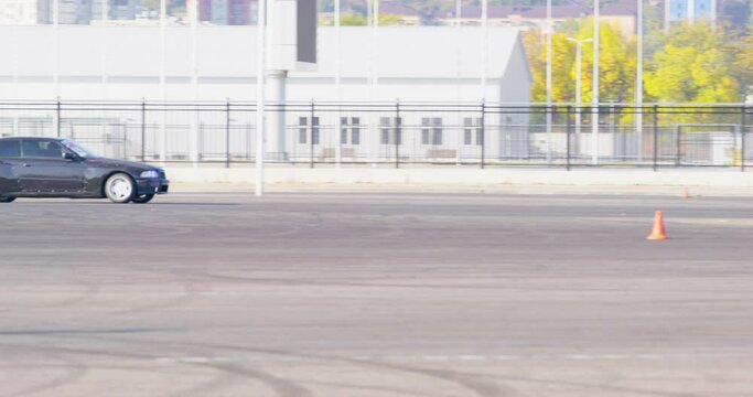 Drifting black car on racing street track fenced from buildings, trees. Vehicle enters turn high speed, judge observes, orange traffic cones on pavement. Automobile sports, entertainment, adrenaline